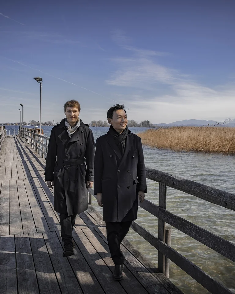 Zwei Männer in dunklen Mänteln (Nils Mönkemeyer und William Youn) spazieren auf einem Holzsteg über einem See mit Schilf und Bergen im Hintergrund.