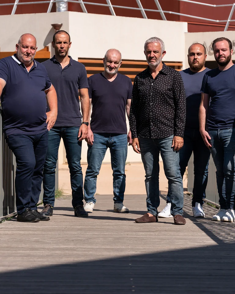 Six men stand on a bridge, wearing casual clothes and smiling at the camera. A modern building is visible in the background.