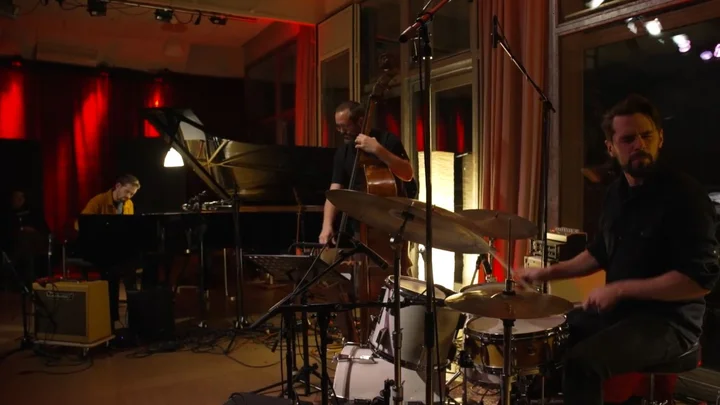 The Pablo Held Trio is playing in a cozy, red-lit room. In the foreground, drummer Jonas Burgwinkel is visible; behind him are bassist Robert Landfermann and pianist Pablo Held.