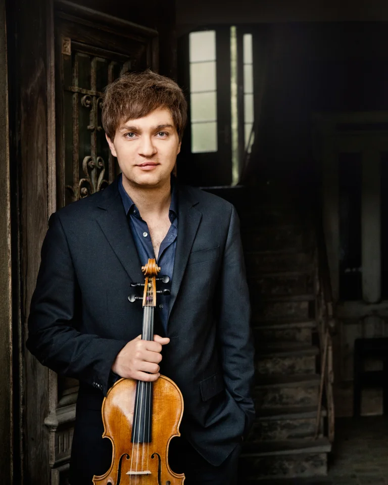Nils Mönkemeyer: A man in a suit stands in a dark room holding a violin. A staircase is visible in the background.