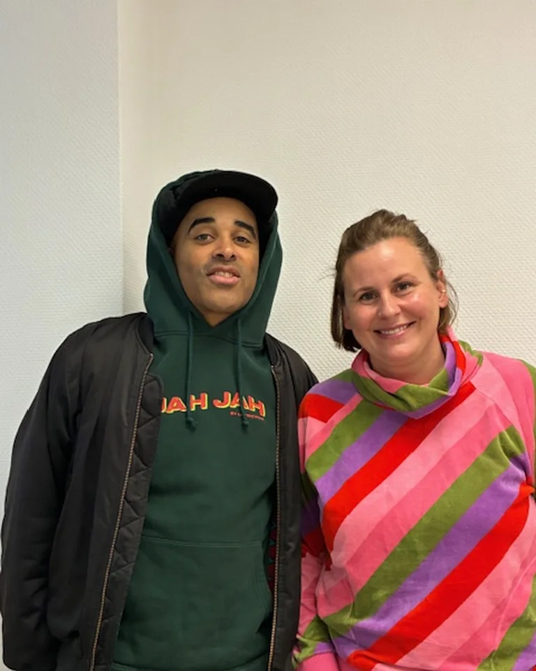 Eine lächelnde Frau in einem bunten Pullover steht neben einem Mann in Kapuzenpullover und Jacke vor einer neutralen Wand.