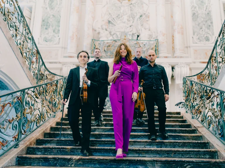 Vier Musiker und eine Frau (Ensemble 1700: Celtic Baroque) in einem lila Anzug gehen eine prächtige Treppe mit kunstvollen Geländern hinunter.