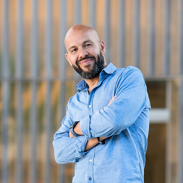 Mourad Merzouki stands in a blue shirt with his arms crossed in front of a blurred wall of vertical stripes.