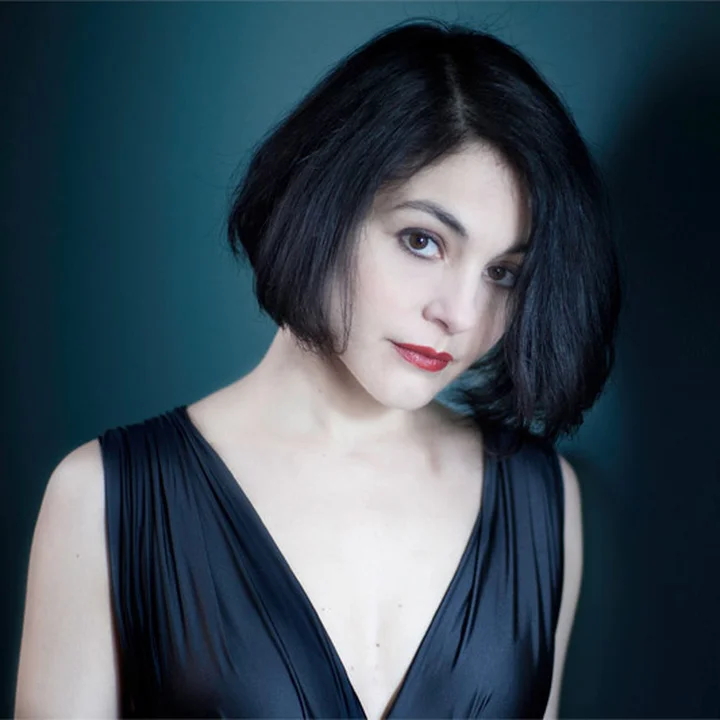 Portrait of Ana Quintans with black, chin-length hair, wearing an elegant, dark dress against a simple blue background.