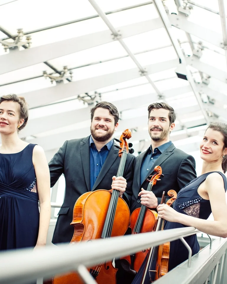 Ein Streichquartett posiert in eleganter Kleidung mit zwei Celli unter einem modernen Glasdach. Die Gruppe lächelt freundlich.