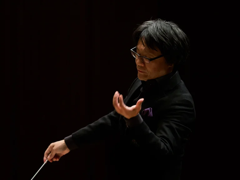 Ein Dirigent (Kiyotaka Teraoka) in dunkler Kleidung leitet mit dramatischen Gesten ein Orchester. Der Hintergrund ist schwarz, wodurch der Fokus auf ihm liegt.
