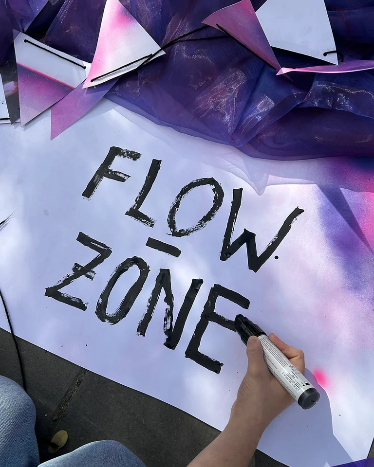 Eine Hand schreibt mit einem schwarzen Marker die Worte »FLOW ZONE« auf ein weißes Blatt Papier mit violetter Dekoration.
