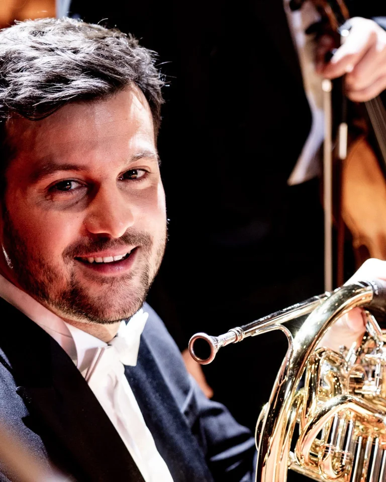 Ein Musiker spielt ein Horn in einem Orchester. Er trägt einen Anzug und lächelt, während er sein Instrument hält.