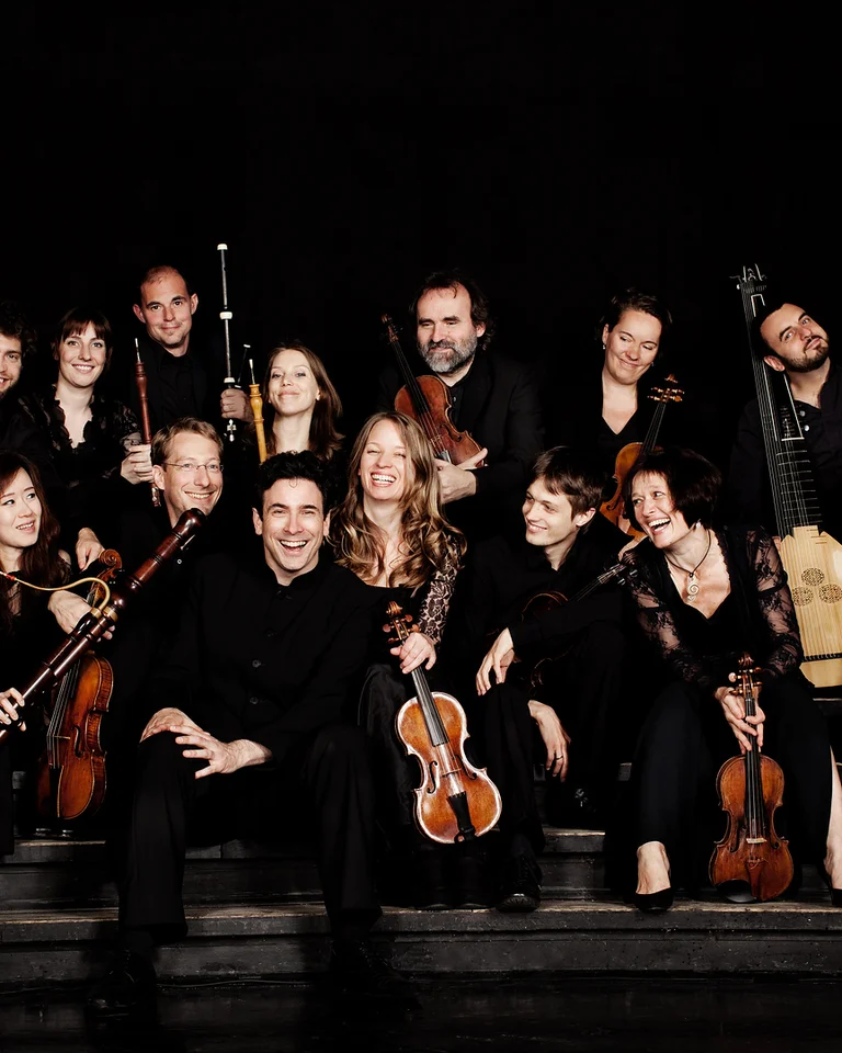 A group of musicians posing with various classical instruments on a dark stage. They are smiling at the camera.