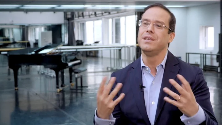 Video still from an interview with Leonardo García-Alarcón, set in a large room with a piano in the background.