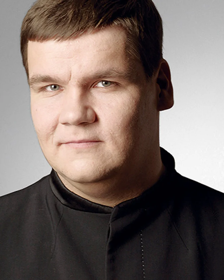 Ein Mann in einem schwarzen Anzug vor einem neutralen grauen Hintergrund. Er hat kurzes, brünettes Haar und schaut ernst.