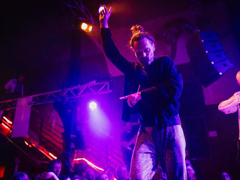 Ein Musiker (aus treppenhausorchester) performt auf einer Bühne mit blau-violetter Beleuchtung. Das Publikum ist im Hintergrund schwach sichtbar. Livemusik-Atmosphäre.