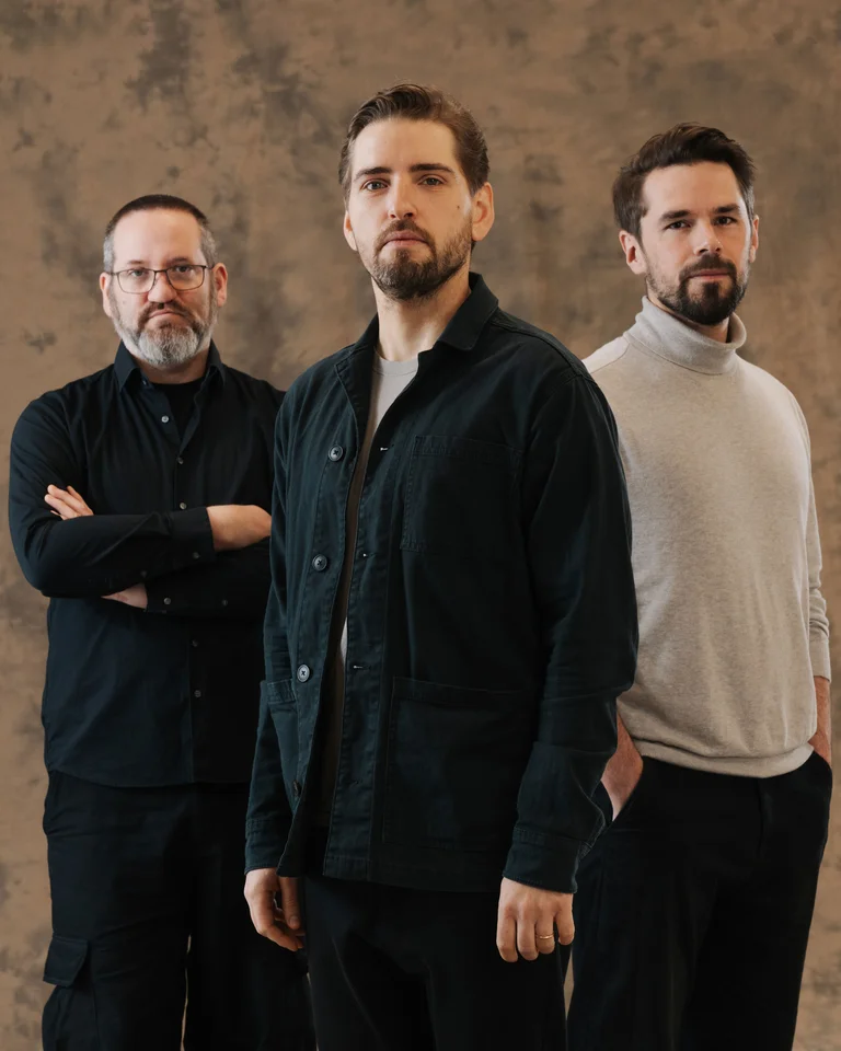 The three members of the Pablo Held Trio stand in dark clothing against a neutral brown background, looking directly at the camera.