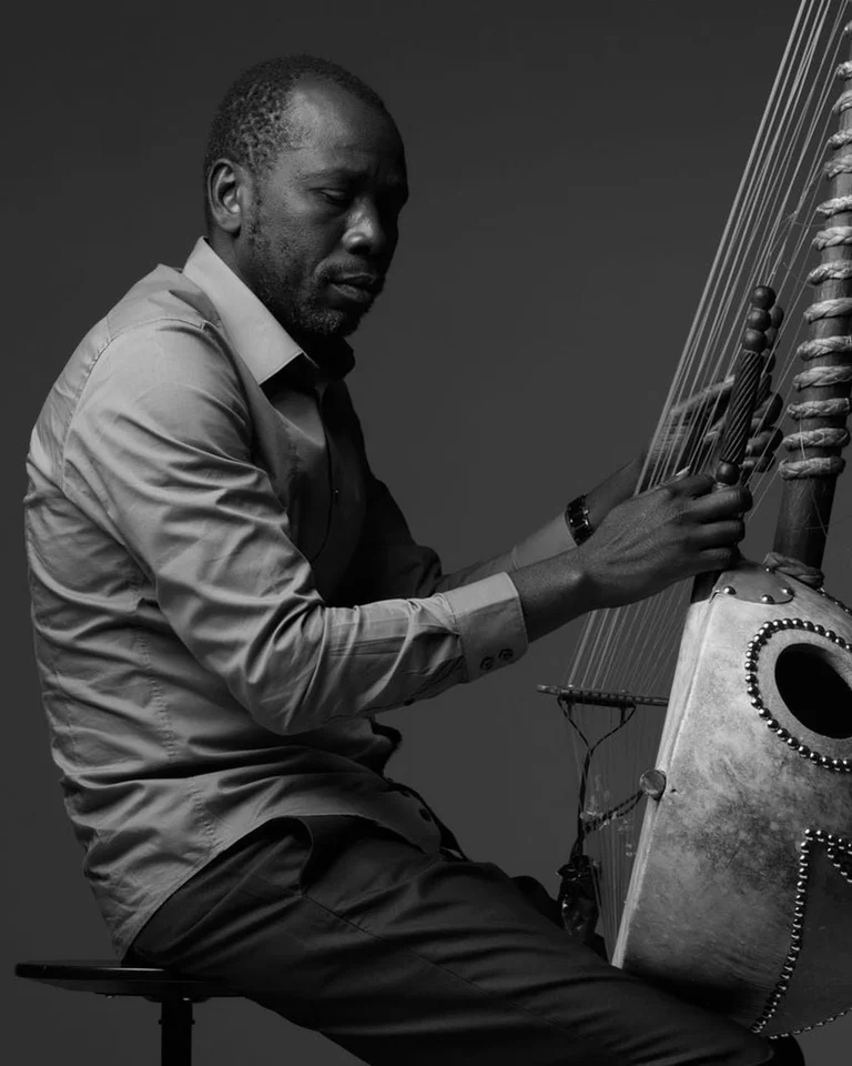 Schwarz-Weiß-Foto eines Mannes (Sissoko Ballaké) , der eine Kora spielt. Er sitzt auf einem Hocker und konzentriert sich auf das Instrument.