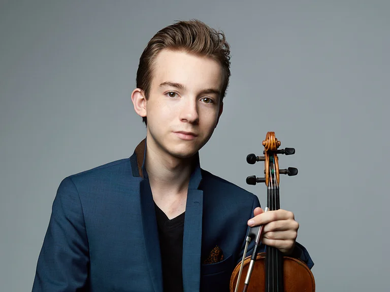 Ein junger Mann (Jacques Forestier) sitzt auf einem Hocker und hält eine Violine. Er trägt eine blaue Jacke und Jeans.