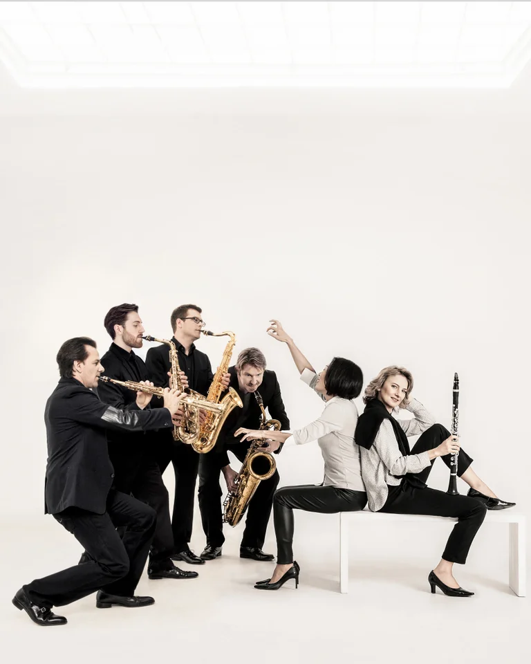 A group of six musicians in suits play various wind instruments in a bright, minimalist room. A woman sits on a bench.