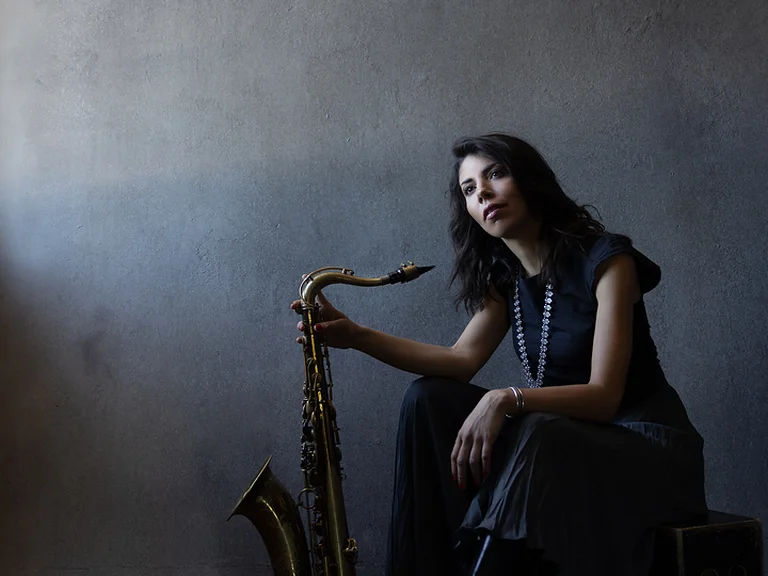 Eine Frau sitzt auf einem Hocker und hält einen Saxophon im Arm. Der Raum ist dunkel und künstlerisch gestaltet.