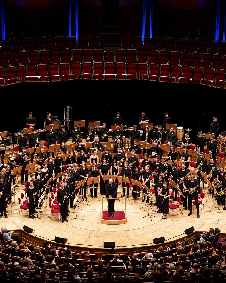 Ein großes Orchester (Symphonisches Jugendblasorchester) spielt in einem gut besuchten Konzertsaal. Der Dirigent steht in der Mitte auf einem Podest.