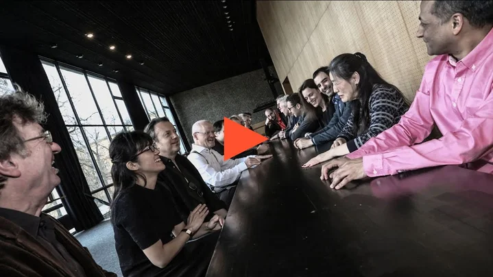 Members of the Ensemble Modern sit at a long table in a modern room with large windows and wood paneling.
