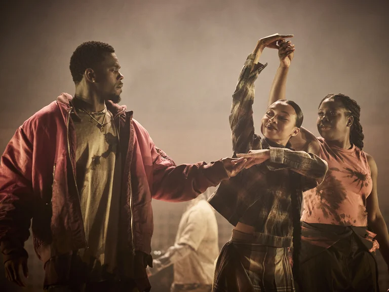 Three dancers (Structure Rualitè) move under dramatic lighting. One raises an arm elegantly, their posture expressive and dynamic.