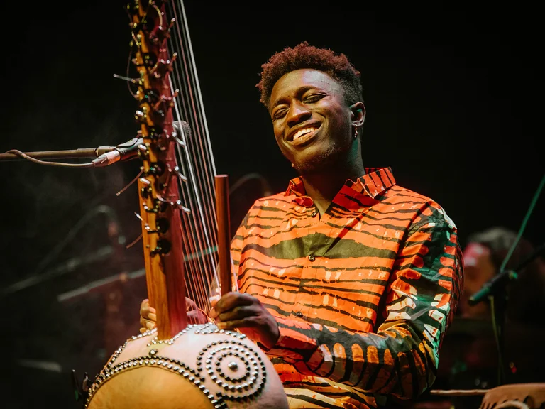 Ein Musiker lächelt, während er eine Kora spielt. Er trägt ein bunt gestreiftes Hemd. Dunkler Hintergrund deutet auf ein Konzert hin.
