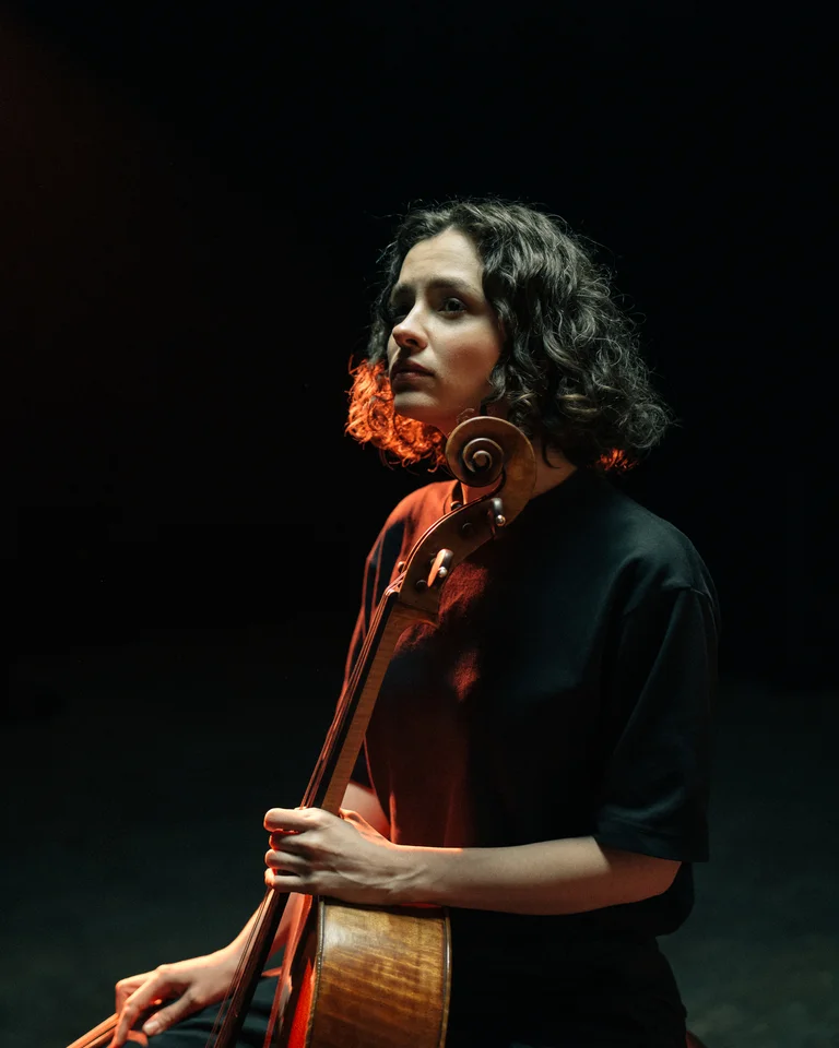 Eine Frau in schwarzer Kleidung spielt in einem dunklen Raum Cello. Das Licht betont ihr Gesicht und das Instrument.