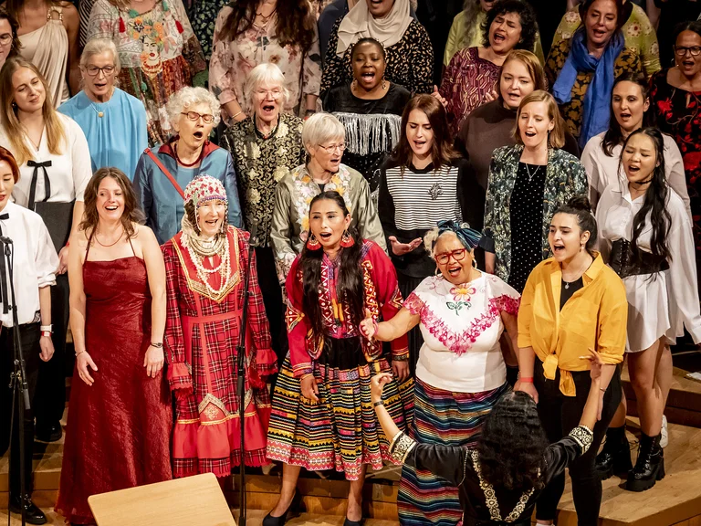 A group of people in colorful traditional clothing stands on a stage, apparently singing together.