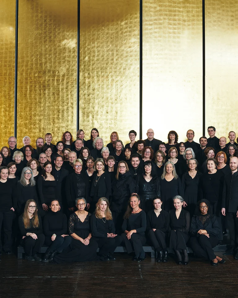 Eine große Gruppe von Menschen in schwarzer Kleidung steht vor einem goldenen Hintergrund. Es handelt sich um einen klassischen Chor.