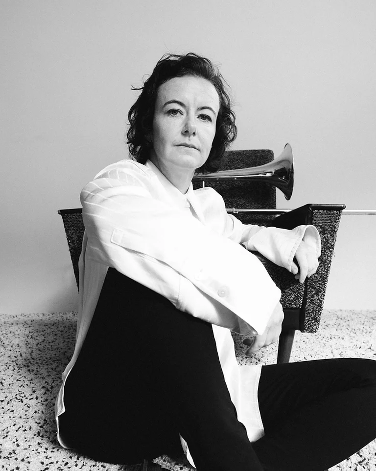 Eine Frau (Shannon Barnett) mit lockigem Haar sitzt in lässiger Pose auf dem Boden vor einer neutralen Kulisse, schwarz-weiß fotografiert.