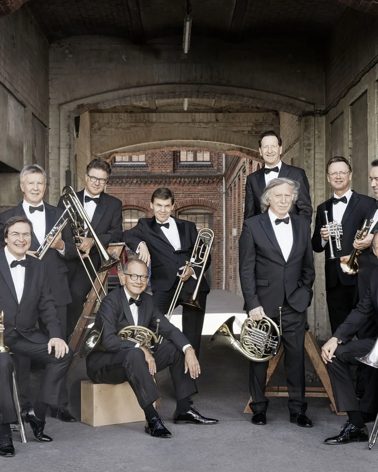 A group of eleven musicians in formal suits poses with brass instruments in front of a brick building. They smile.