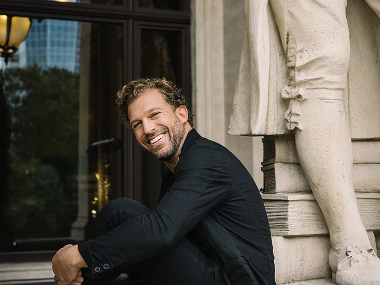 Ein Mann (Julian Prégardien) sitzt lächelnd auf einem Gebäudevorsprung neben einer Statue. Im Hintergrund sind Glasgebäude zu sehen.