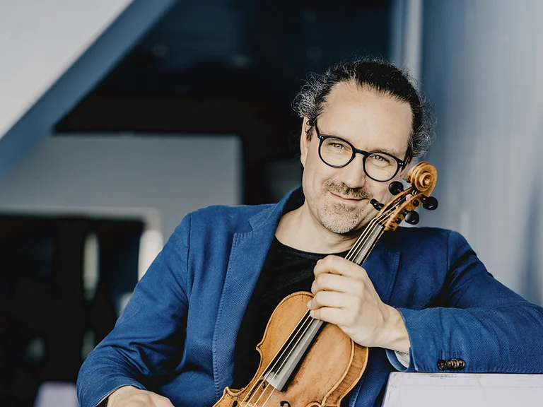 Mann (Julien Chauvin) mit Brille und blauer Jacke sitzt und hält eine Geige. Er lächelt leicht und wirkt entspannt.