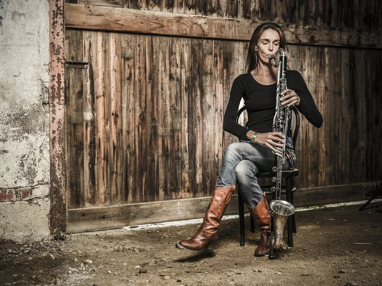 Eine Frau (Tara Bouman) sitzt auf einem Stuhl vor einer rustikalen Holzwand und spielt Klarinette. Sie trägt Jeans und braune Stiefel.