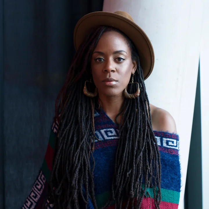 Portrait of Akua Naru with long dreadlocks, a hat, and a colorful, patterned top. She is standing against a dark background.