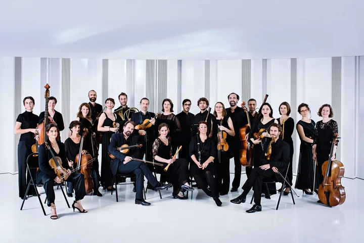 The members of the orchestra Le Concert de la Loge pose in elegant black attire, holding various instruments, against a white background.