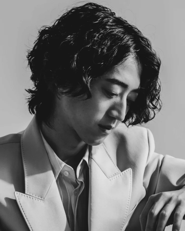 Ein Schwarz-Weiß-Foto einer Person mit lockigem Haar und Anzug. Sie sitzt nachdenklich neben einem Klavier.