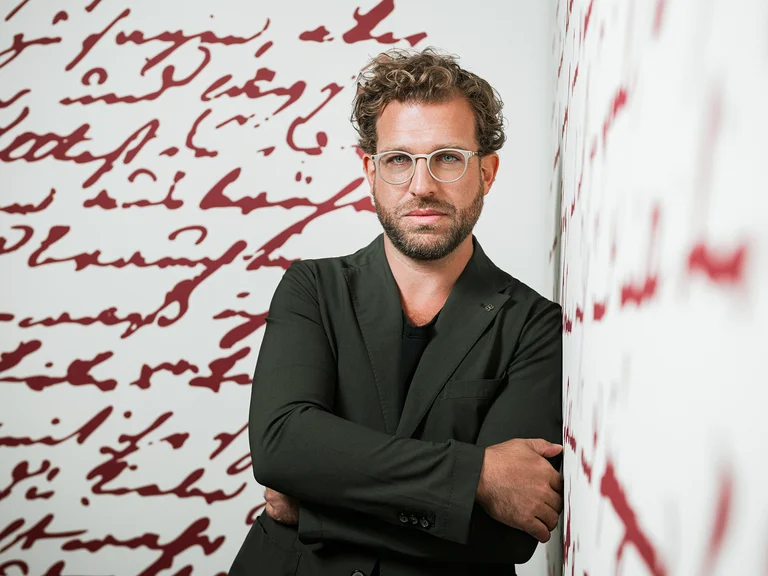 Eine Person (Julian Prégardien) mit Brille steht vor einer Wand mit roter, handgeschriebener Schrift. Sie trägt einen schwarzen Anzug.