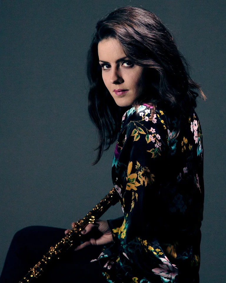 Eine Frau mit langen dunklen Haaren hält eine Oboe. Sie trägt ein gemustertes Kleid vor einem dunklen Hintergrund.