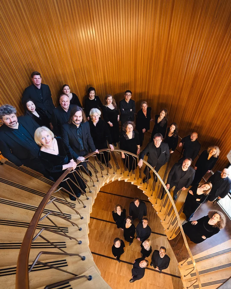 Eine Gruppe von Menschen steht auf einer spiralförmigen Treppe aus Holz. Sie tragen schwarze Kleidung und schauen nach oben.