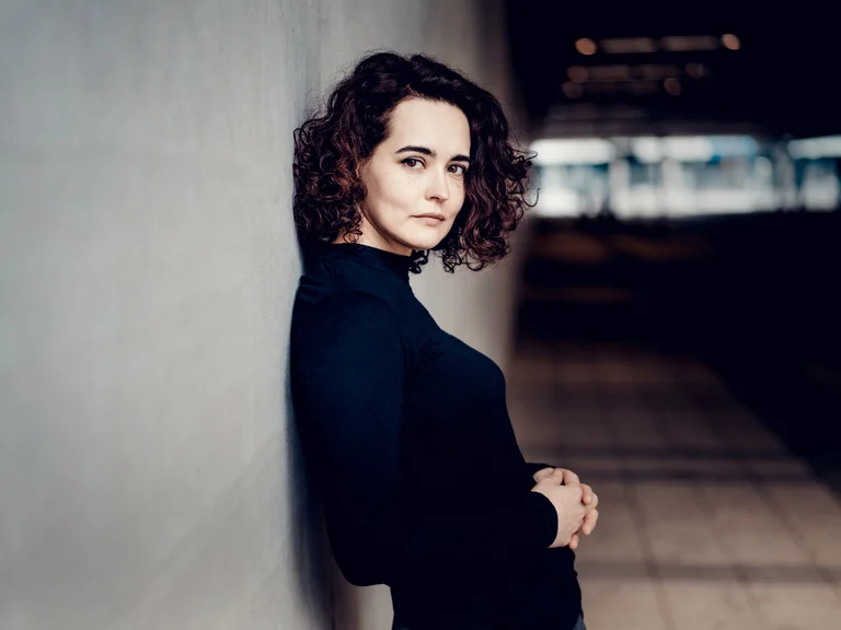 Alena Baeva: Eine Frau mit lockigem Haar lehnt an einer Wand. Sie trägt einen schwarzen Pullover und schaut selbstbewusst in die Kamera.