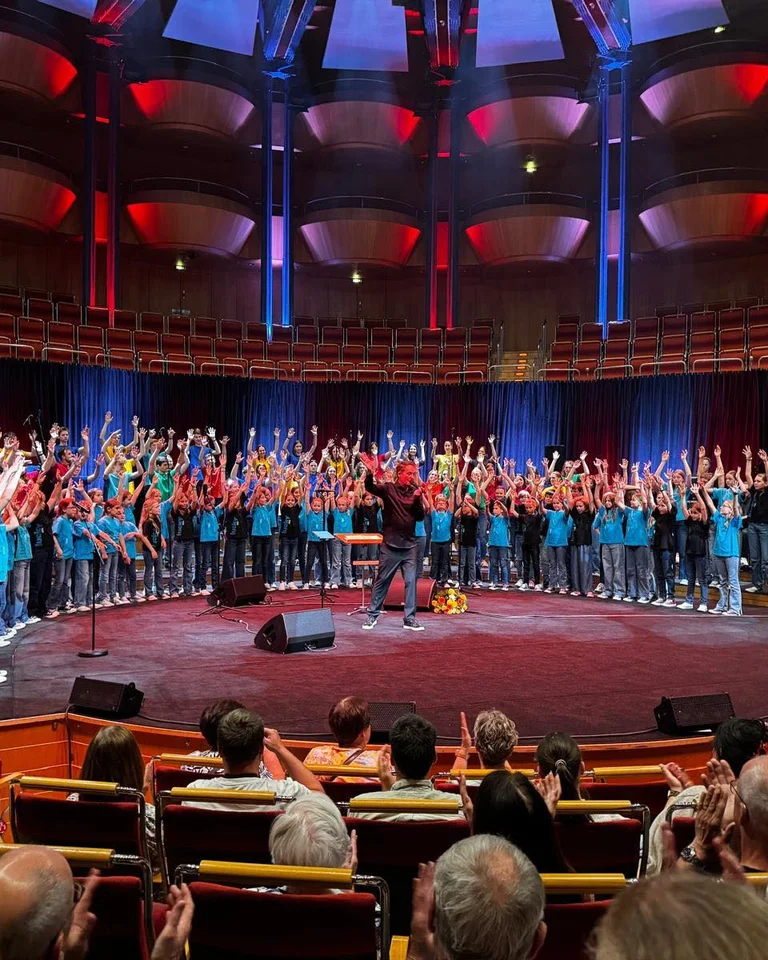 Kölner Jugendchor St.  Stephan & Lucky Kids: Ein großer Chor singt auf einer beleuchteten Bühne in einem Konzertsaal. Das Publikum applaudiert begeistert.