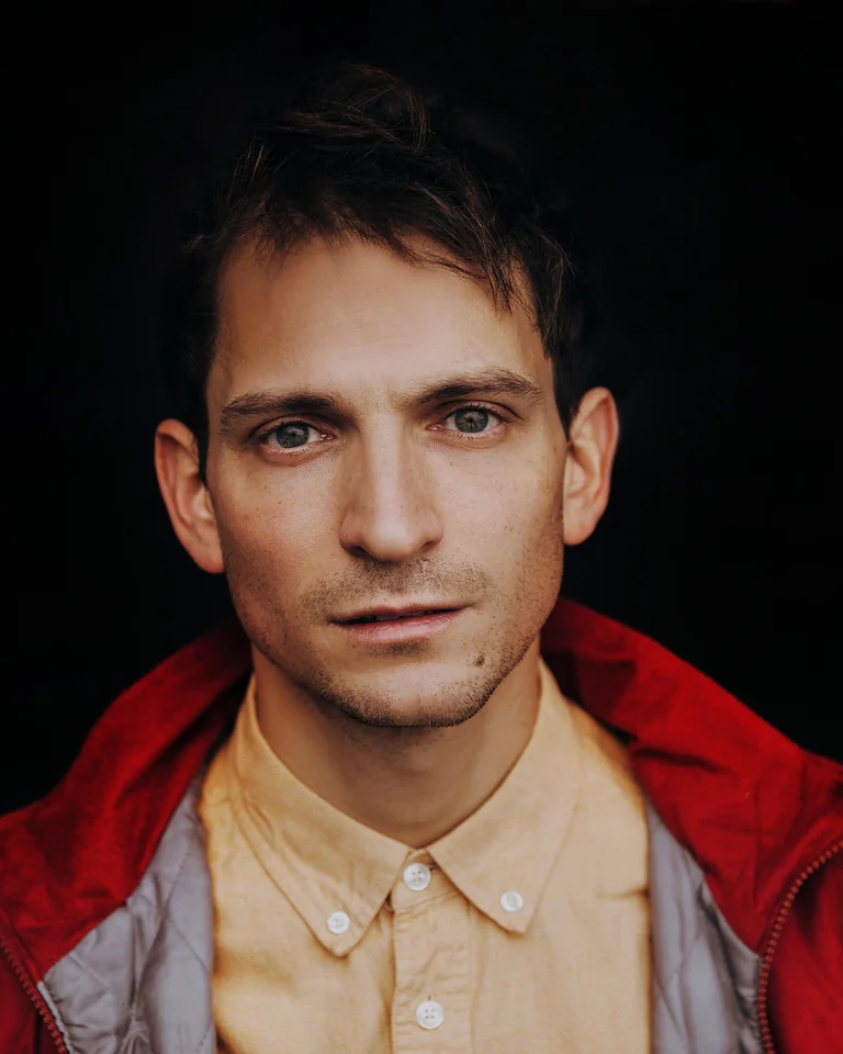 Ein Mann mit kurzem dunklem Haar trägt eine rote Jacke über einem beigen Hemd. Er blickt direkt in die Kamera.