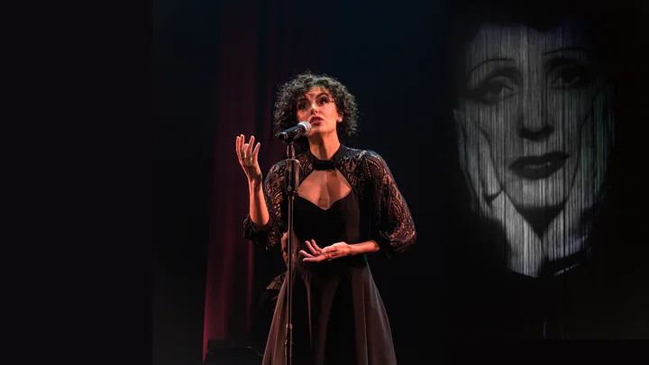 Eine Frau singt auf einer Bühne, im Hintergrund ein großes schwarz-weiß Bild von Edith Piaf. Emotionale Darbietung.