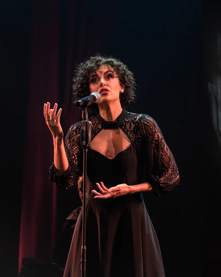 Eine Frau singt auf einer Bühne, im Hintergrund ein großes schwarz-weiß Bild von Edith Piaf. Emotionale Darbietung.