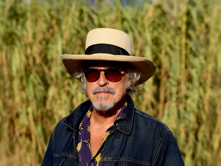 Ein Mann mit Strohhut und Sonnenbrille steht vor einem Feld aus hohen Pflanzen. Er trägt eine dunkle Jacke.