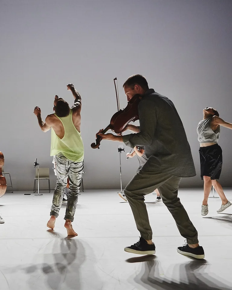 Tänzer und Musiker mit Streichinstrumenten bewegen sich dynamisch auf einer Bühne. (Scottish Ensemble) Die Szene strahlt Energie und Kreativität aus.