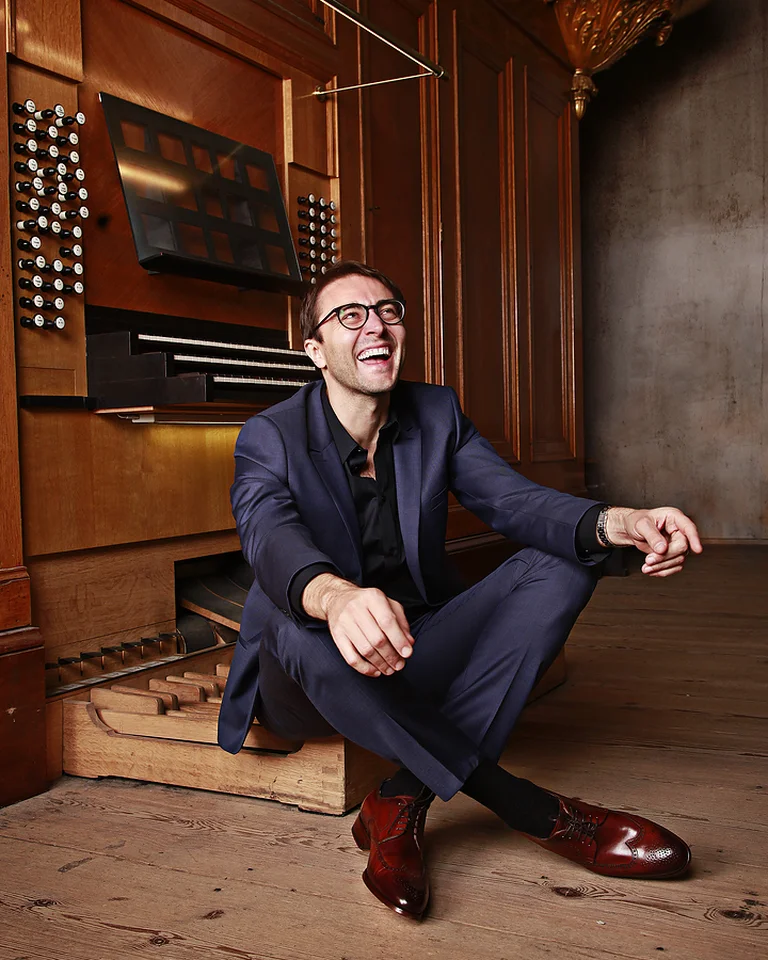 A man in a suit (Christian Schmitt) sits laughing on the floor in front of a large organ in an elegant, wood-paneled room.
