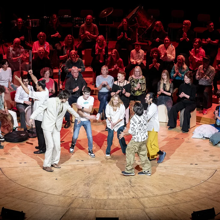 A colorful group performs on stage in front of a seated audience. The spectators are attentive and smiling.
