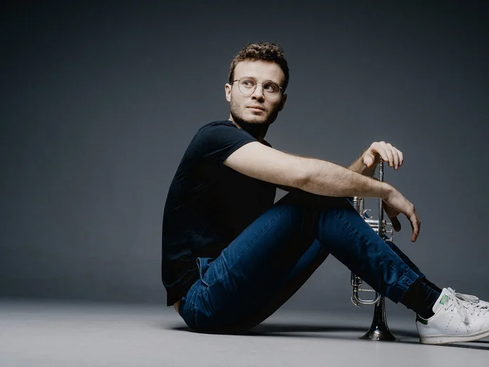 Eine Person (Simon Höfele) in Jeans und T-Shirt sitzt auf dem Boden, mit einer dunklen, minimalistischen Kulisse im Hintergrund.