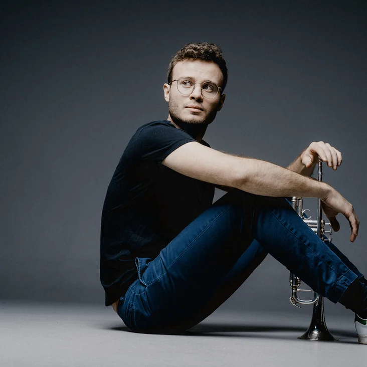 Eine Person (Simon Höfele) in Jeans und T-Shirt sitzt auf dem Boden, mit einer dunklen, minimalistischen Kulisse im Hintergrund.